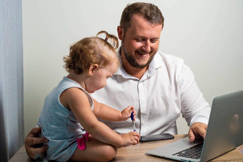 Happy dad working at home with a laptop, holding the baby in his arms